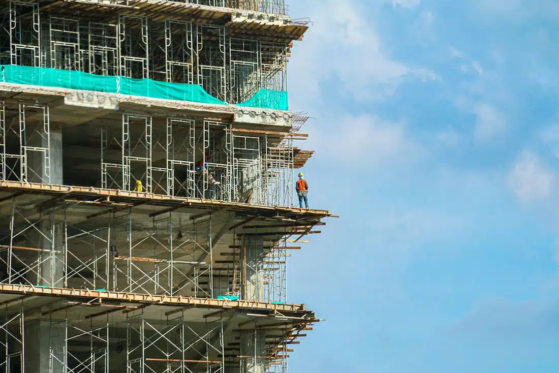 The corner of a building under heavy construction with a worker standing at the building’s edge