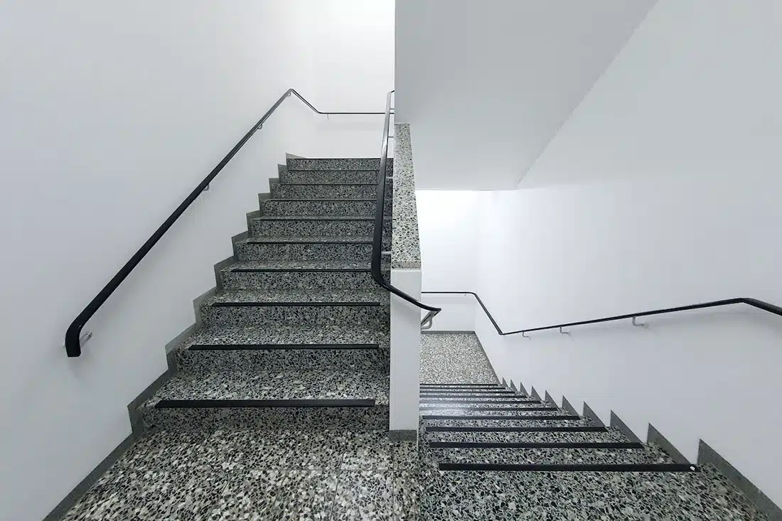 A shot from a landing of a white stairwell. Grey-speckled stairs go upwards to the left and down to the right