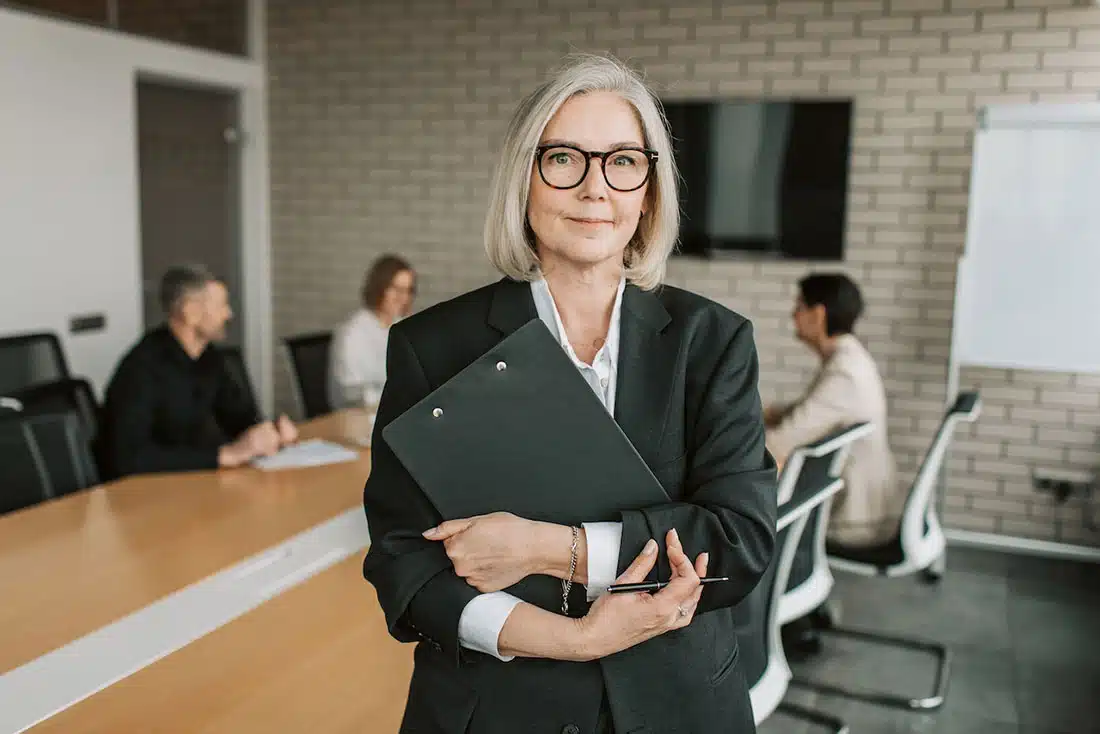 A woman with short white hair in a black blazer holding a clipboard. Three people in the background are holding a meeting
