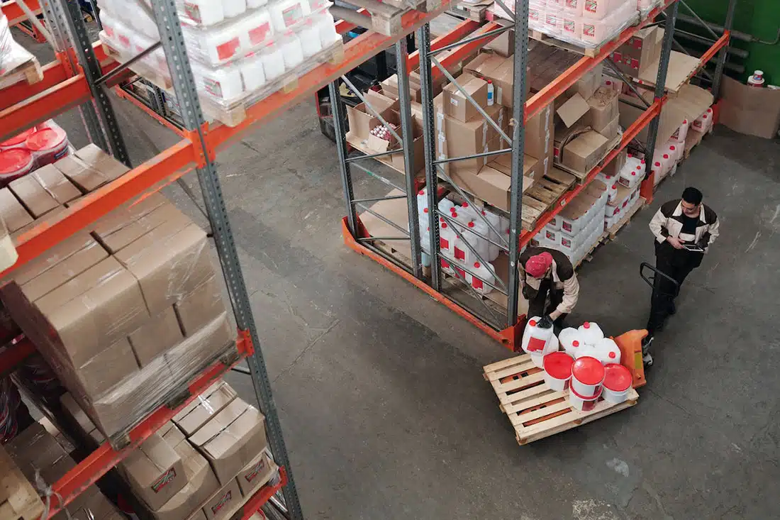 A bird’s eye view of two factory workers adding large jugs of liquid to a lower shelf. There are rows of boxes on higher shelves