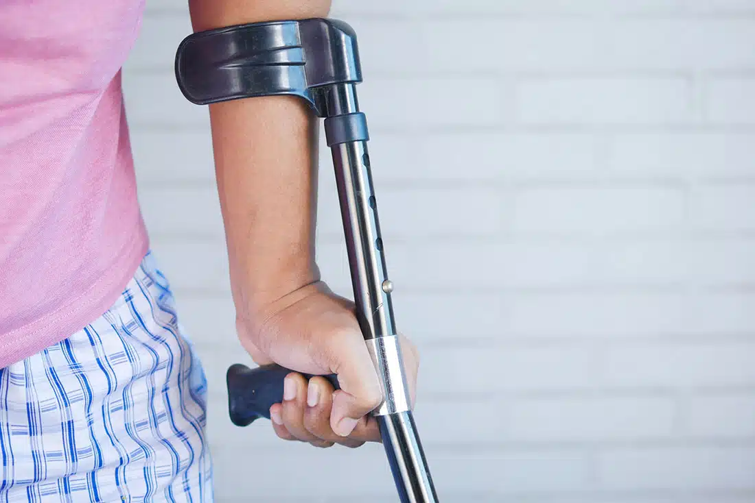 “A close-up shot of someone’s torso and left arm. They have a crutch. They’re wearing a pink tee and blue checked shorts