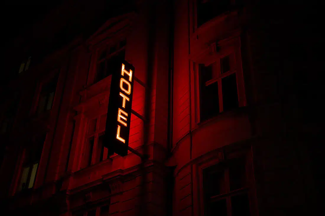 A close-up of the side of a Victorian building in the dark. It has a red neon sign that says ‘hotel,’ between windows